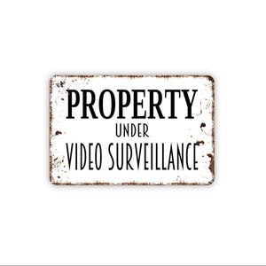May include: A rectangular metal sign with a white background and distressed brown edges. The text in black, sans-serif font reads PROPERTY UNDER VIDEO SURVEILLANCE. The sign features a weathered, vintage-style finish with rust-like markings.