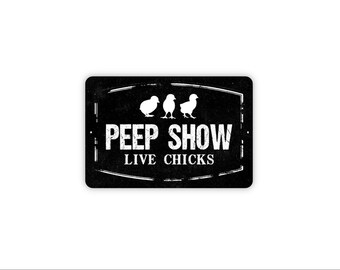 May include: A rectangular black metal sign with a distressed white border. The sign features three white silhouettes of baby chicks above the white text PEEP SHOW and the text LIVE CHICKS in a smaller font size below the main heading.