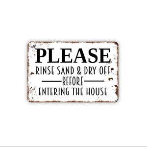 May include: A rectangular white metal sign with distressed brown edges featuring the black text PLEASE RINSE SAND & DRY OFF BEFORE ENTERING THE HOUSE. The sign has a weathered appearance with rust-like markings along the border and corners.