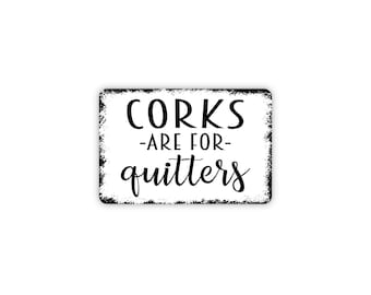 May include: A rectangular white metal sign with rounded corners and a distressed black border. The sign features black text that reads CORKS -ARE FOR- quitters in a combination of bold block lettering and a decorative cursive script font.