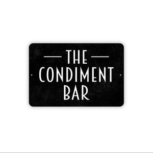 May include: A rectangular black sign with rounded corners featuring white text that reads THE CONDIMENT BAR. The design includes a horizontal line above the word THE and a horizontal line below the word BAR, with small mounting holes on the sides.