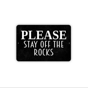 May include: A rectangular black sign with rounded corners featuring the white text PLEASE STAY OFF THE ROCKS in a clean, sans-serif font. The sign has a subtle textured finish and two small mounting holes on the left and right sides.