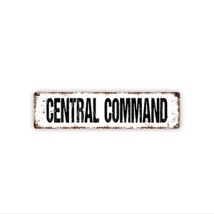 May include: A rectangular metal sign with a white background and a distressed, rusted border. The text CENTRAL COMMAND is printed in bold, black, sans-serif capital letters across the center of the sign, featuring a weathered, worn appearance.