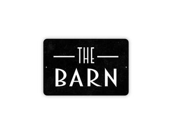May include: A rectangular black metal sign with rounded corners featuring the white text THE BARN in a stylized sans-serif font. The word THE is centered above the word BARN, with a horizontal white line on each side of the word THE.