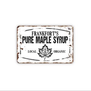 May include: A rectangular metal sign with a distressed white finish and rusted edges. The text reads FRANKFORT'S PURE MAPLE SYRUP with the words LOCAL and ORGANIC on either side of a central black maple leaf illustration, all enclosed in a border.