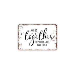 May include: A rectangular metal sign with a white background and distressed brown edges. The text reads AND SO together THEY BUILT A LIFE THEY LOVED in a combination of black script and sans-serif fonts, centered on the surface of the sign.