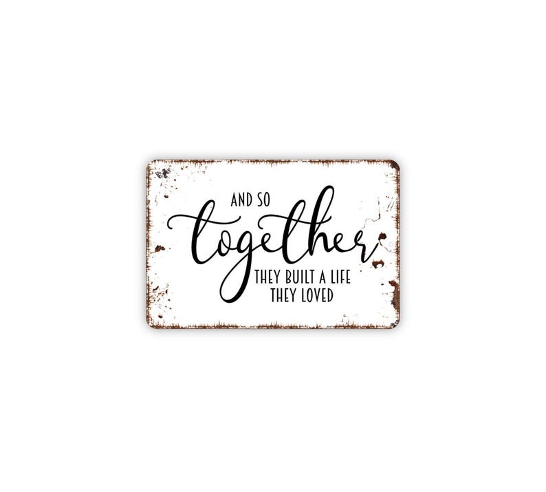 May include: A rectangular metal sign with a white background and distressed brown edges. The text reads AND SO together THEY BUILT A LIFE THEY LOVED in a combination of black script and sans-serif fonts, centered on the surface of the sign.