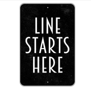 May include: A rectangular black sign with rounded corners and a subtle textured finish. The white text in a clean, sans-serif font reads LINE STARTS HERE in three stacked lines. There are two small mounting holes at the top and bottom center.
