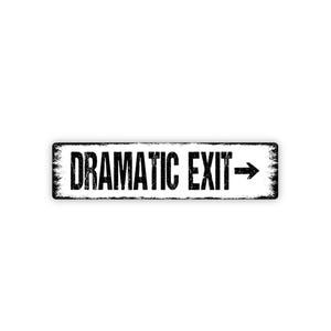 May include: A rectangular black and white sign with the words "DRAMATIC EXIT" in bold, distressed font. An arrow points to the right. The sign has a weathered, vintage appearance, suggesting it's a decorative item.