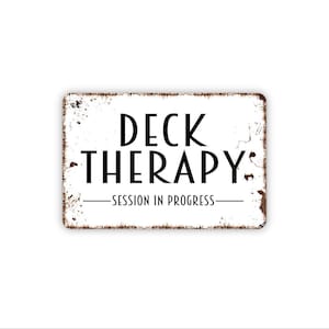 May include: A rectangular white metal sign with rounded corners and distressed brown edges. The text DECK THERAPY is printed in large black letters, followed by the text SESSION IN PROGRESS centered between two horizontal black lines.