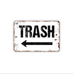 May include: A rectangular metal sign with a white background and distressed brown edges. The sign features the word TRASH in bold, black, distressed capital letters, positioned above a black arrow pointing to the left on a white surface.