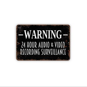 May include: A rectangular black metal sign with a distressed, rusted edge. The text in white reads: WARNING - 24 HOUR AUDIO & VIDEO RECORDING SURVEILLANCE. The sign features a bold, serif font for the word WARNING and a clean, sans-serif font below.