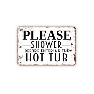 May include: A rectangular white metal sign with rounded corners and a distressed, rusted edge. The sign features black text that reads: PLEASE SHOWER BEFORE ENTERING THE HOT TUB, with a horizontal line separating the words PLEASE and SHOWER.