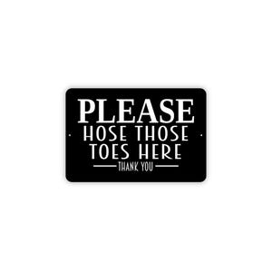 May include: A rectangular black sign with rounded corners featuring white text that reads PLEASE HOSE THOSE TOES HERE followed by THANK YOU with horizontal lines on either side. The sign has small mounting holes on the left and right sides.