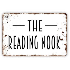 May include: A rectangular white metal sign with rounded corners and a distressed, rusted edge. The sign features black text that reads THE READING NOOK, with a horizontal line centered above the word THE. The design has a weathered appearance.
