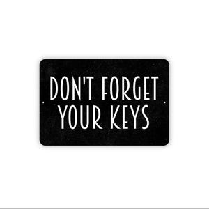 May include: A rectangular black metal sign with rounded corners featuring the white text DON'T FORGET YOUR KEYS in a clean sans-serif font. The sign has two small mounting holes on the left and right sides, centered vertically on the edges.