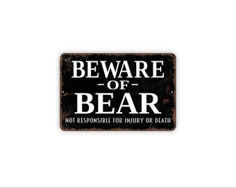 May include: A rectangular black metal sign with a distressed, rusted edge effect. The sign features white text that reads BEWARE OF BEAR in large capital letters, with the smaller text NOT RESPONSIBLE FOR INJURY OR DEATH printed below.