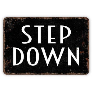 May include: A rectangular metal sign with a black background and white text that reads STEP DOWN. The sign features a distressed, weathered finish with visible rust-colored edges and small mounting holes on the left and right sides.