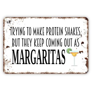 May include: A rectangular metal sign with a distressed white finish and brown rusted edges. The black text reads: TRYING TO MAKE PROTEIN SHAKES, BUT THEY KEEP COMING OUT AS MARGARITAS. A small illustration of a margarita with a lime wedge is included.