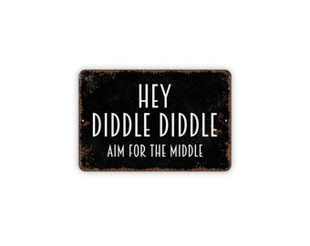 Peut inclure: Une plaque rectangulaire en métal noir avec des bords rouillés et usés. Le texte en police blanche sans empattement indique : HEY DIDDLE DIDDLE AIM FOR THE MIDDLE. La plaque présente de petits trous de fixation sur les côtés gauche et droit.