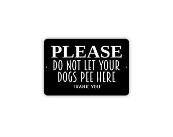 Op de afbeelding: Een rechthoekig zwart bord met afgeronde hoeken en witte tekst die luidt: PLEASE DO NOT LET YOUR DOGS PEE HERE THANK YOU. Het bord heeft twee kleine ronde gaten aan de linker- en rechterkant voor bevestigingsdoeleinden.