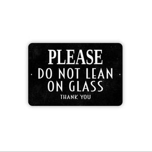 May include: A rectangular black sign with rounded corners featuring white text that reads PLEASE DO NOT LEAN ON GLASS THANK YOU. The sign has a matte finish and a simple, clean design with clear, legible lettering centered on the surface.