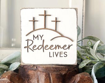 May include: White wooden sign with the text "My Redeemer Lives" and three wooden crosses on a hill. The sign is sitting on a wooden stand.