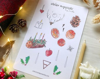 Può includere: Una carta bianca con illustrazioni ad acquerello di elementi festivi, tra cui candele, una testa di cervo, fette d'arancia e agrifoglio. La carta è circondata da pigne, una coperta a quadri rossa e nera e luci fatate. Il testo "atelier myosotis" è visibile.
