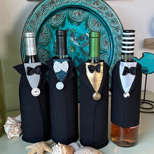 May include: Four wine bottles dressed in miniature tuxedo jackets with bow ties and buttons. The jackets are black, with silver, white, and gold accents. The bottles are arranged on a white surface with seashells and a starfish.