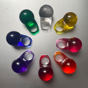 以下が含まれることがあります： カラフルな樹脂リングのコレクション。各リングは、バンドの上に丸く半透明のオーブが特徴です。リングは、緑、クリア、黄色、ピンク、オレンジ、赤、紫、青など、さまざまな色があります。各リングには「MIR」の文字がプリントされています。