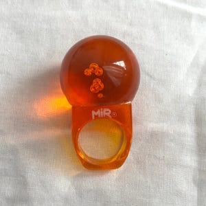 以下が含まれることがあります： 大きな丸い泡の入ったトップが付いたオレンジ色の樹脂リング。 リングバンドは長方形で、前面に「MIR」の文字がプリントされています。 リングは鮮やかなオレンジ色です。