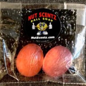 Puede incluir: Dos pequeñas bolas de jabón redondas de color naranja en una bolsa de plástico transparente. La bolsa tiene una pegatina con el texto "Nut Scentz Ball Soap" y un personaje de dibujos animados con zapatillas.