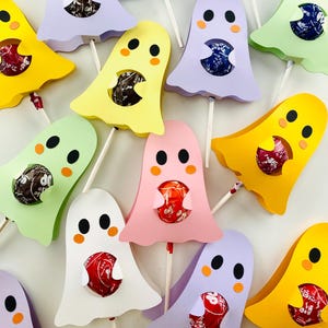 Pode incluir: Uma coleção de suportes de pirulitos em forma de fantasma em várias cores pastel, incluindo rosa, amarelo, azul e verde. Cada suporte tem um doce redondo visível no centro e é preso a um palito de madeira. Os rostos dos fantasmas têm olhos pretos e bochechas laranja.