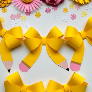 Może przedstawiać: Trzy żółte papierowe kokardki z końcówkami w kształcie ołówka. Kokardki są ułożone w rzędzie, dwie górne zwrócone do siebie, a dolna skierowana w dół. Kokardki znajdują się na białym tle.