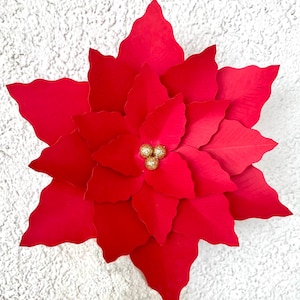 以下が含まれることがあります： 金色のグリッターのアクセントが付いた、大きな赤い紙のポインセチアの花。花は複数の紙の層でできており、3 次元的な外観をしています。