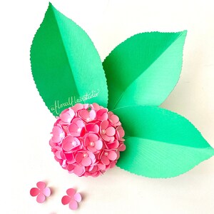 Peut inclure: Une fleur en papier rose avec un centre blanc, entour&eacute;e de trois feuilles en papier vert. La fleur est compos&eacute;e de nombreuses petites p&eacute;tales en papier et a un aspect textur&eacute;. Les feuilles sont &eacute;galement textur&eacute;es et ont un bord festonn&eacute;.