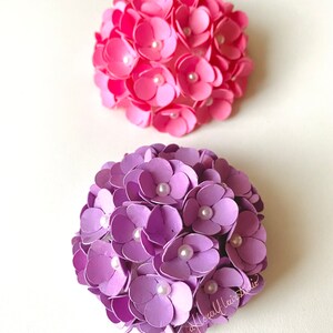 Peut inclure: Deux arrangements de fleurs en papier, l'un rose et l'autre violet, avec des accents de perles blanches. Les fleurs sont dispos&eacute;es en forme de sph&egrave;re.