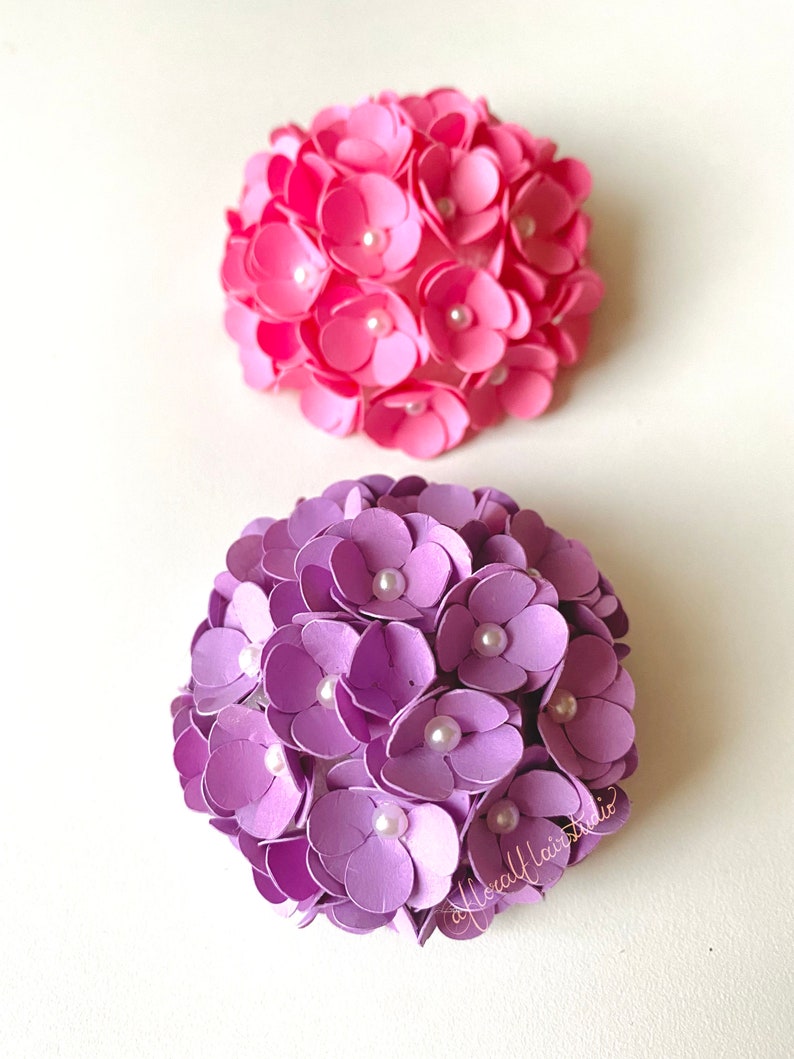 Peut inclure: Deux arrangements de fleurs en papier, l'un rose et l'autre violet, avec des accents de perles blanches. Les fleurs sont dispos&eacute;es en forme de sph&egrave;re.