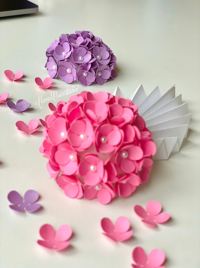 Peut inclure: Deux arrangements de fleurs en papier, l'un rose et l'autre violet, avec des accents de perles blanches. Les fleurs sont dispos&eacute;es en forme de sph&egrave;re et sont entour&eacute;es de p&eacute;tales de fleurs en papier d&eacute;tach&eacute;s.