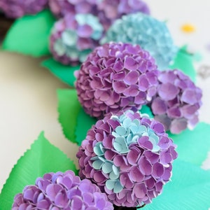 Peut inclure: Une rang&eacute;e d'hortensias en papier dans des tons de violet et de bleu, avec des feuilles vertes. Les hortensias sont faits de p&eacute;tales de papier individuels.