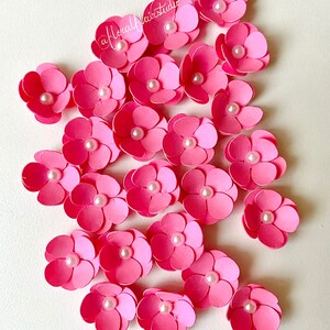 Peut inclure: Un ensemble de 20 fleurs en papier rose avec des centres de perles blanches. Les fleurs sont dispos&eacute;es en grappe sur un fond blanc.