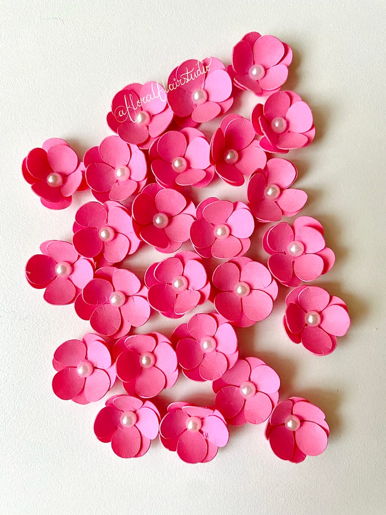 Peut inclure: Un ensemble de 20 fleurs en papier rose avec des centres de perles blanches. Les fleurs sont dispos&eacute;es en grappe sur un fond blanc.
