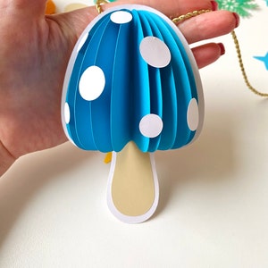 May include: A blue paper mushroom ornament with white polka dots and a beige stem. The ornament is hanging from a gold string.