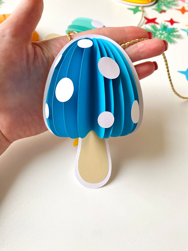 May include: A blue paper mushroom ornament with white polka dots and a beige stem. The ornament is hanging from a gold string.