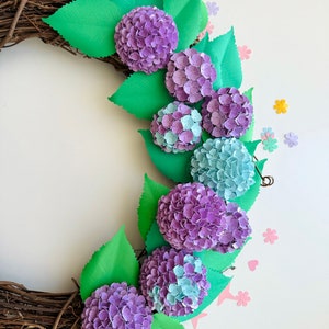 Peut inclure: Une couronne de fleurs en papier avec des hortensias violets et bleus et des feuilles vertes. La couronne est faite de brindilles brunes et est d&eacute;cor&eacute;e de petites fleurs en papier de diff&eacute;rentes couleurs.