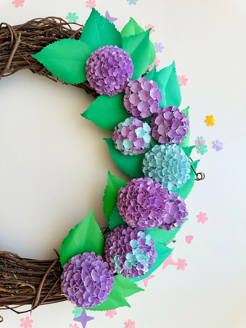 Peut inclure: Une couronne de fleurs en papier avec des hortensias violets et bleus et des feuilles vertes. La couronne est faite de brindilles brunes et est d&eacute;cor&eacute;e de petites fleurs en papier de diff&eacute;rentes couleurs.