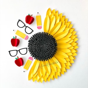 May include: A paper craft sunflower with a black center, surrounded by yellow petals.  There are three red apples, three yellow pencils, and two pairs of black glasses arranged around the sunflower. The text "@floralfairstudio" is written in green cursive.