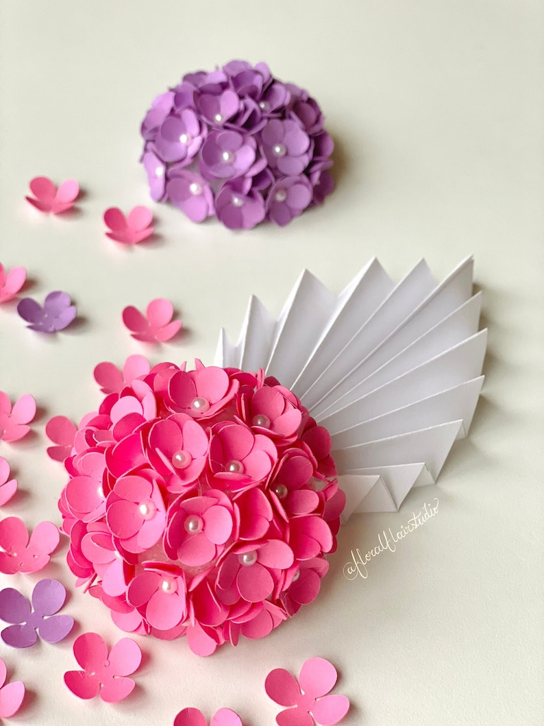 Peut inclure: Deux arrangements de fleurs en papier, l'un rose et l'autre violet, avec des accents de perles blanches. Les fleurs sont dispos&eacute;es en forme de sph&egrave;re et sont entour&eacute;es de plus petites fleurs en papier.