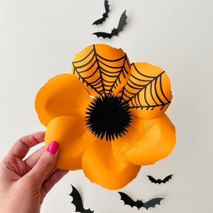 K&ouml;nnte beinhalten: Eine gro&szlig;e orangefarbene Papierblume mit einem schwarzen Spinnennetz-Design und einer schwarzen, flauschigen Mitte. Die Blume wird in einer Hand vor einem wei&szlig;en Hintergrund gehalten, auf dem schwarze Papierflederm&auml;use verstreut sind.