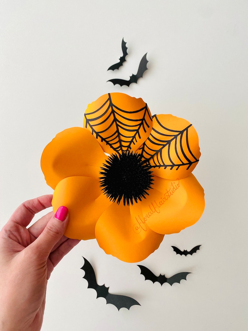 K&ouml;nnte beinhalten: Eine gro&szlig;e orangefarbene Papierblume mit einem schwarzen Spinnennetz-Design und einer schwarzen, flauschigen Mitte. Die Blume wird in einer Hand vor einem wei&szlig;en Hintergrund gehalten, auf dem schwarze Papierflederm&auml;use verstreut sind.